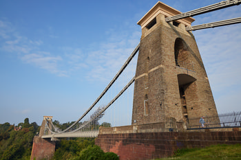 Bristol Suspension Bridge The image shows the Bristol Suspension Bridge in Bristol, England, United Kingdom, captured in the morning during late summer. This architecture photograph prominently features one of the stone towers and the suspension cables of the bridge, spanning across the Avon Gorge with lush green vegetation below. The Bristol Suspension Bridge is an iconic landmark recognized for its impressive engineering and design, and this view highlights its structural details as well as its integration with the surrounding landscape in Bristol.